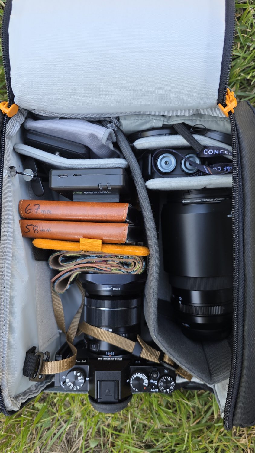 An open Lowepro backpack on the grass, packed with the camera, a long lens, filters, and accessories.
