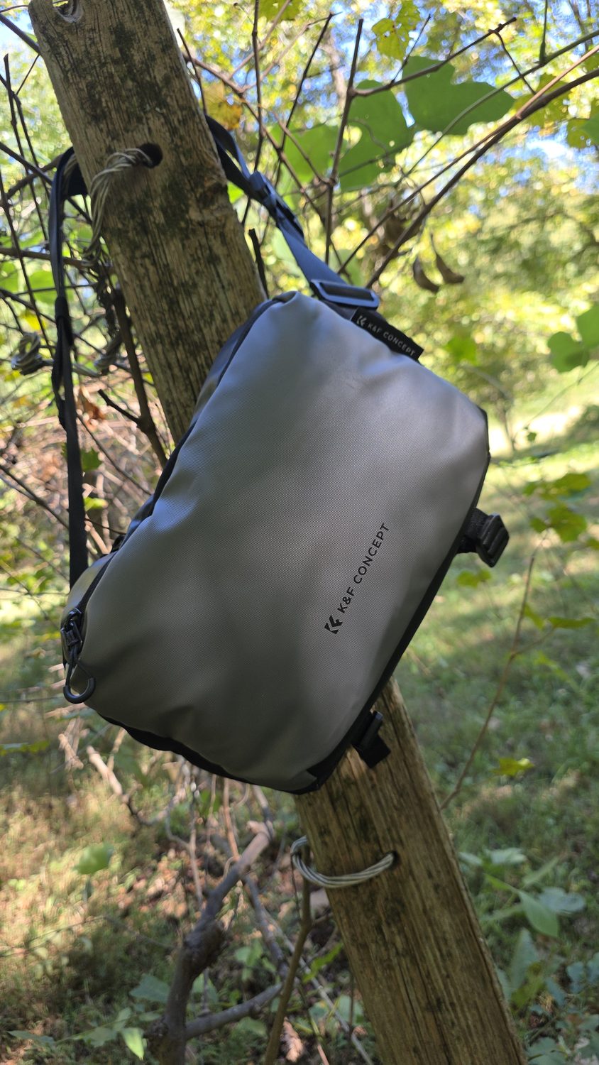 A K&F Concept sling bag hanging from a post in the woods.