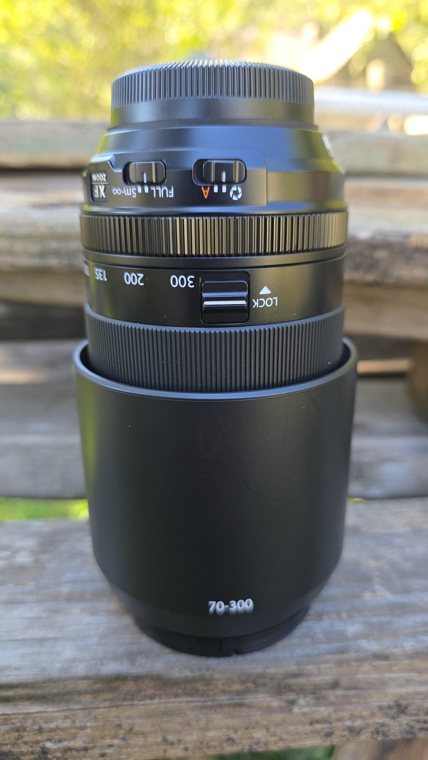 The XF70-300mm lens standing upright on a wooden rail, hood attached, with its focus and stabilizer switches on the side.