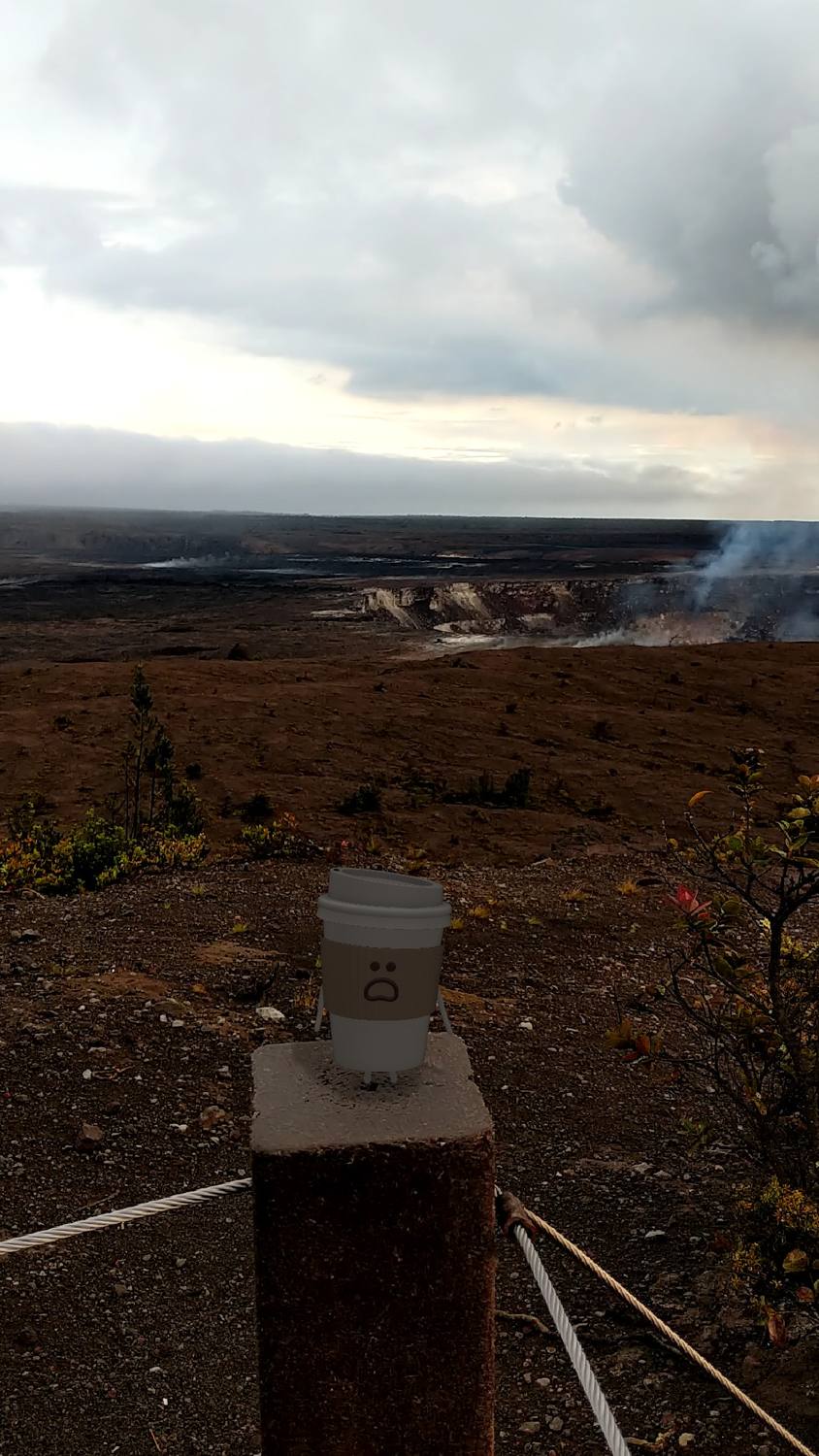 A paper coffee cup sitting on a wooden railing post, overlooking a dark volcanic plain and a distant steaming crater.