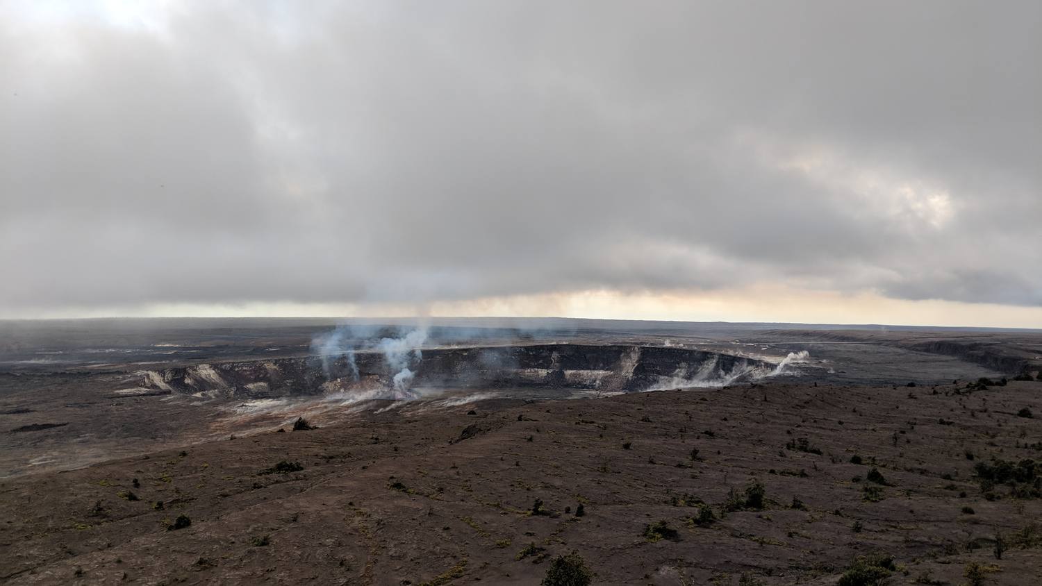A wide, flat volcanic landscape under heavy gray clouds, with white steam rising from a crater in the distance.