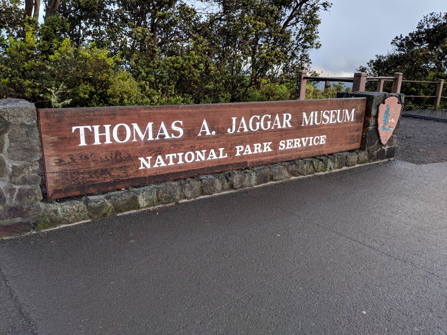 A long wooden sign reading Thomas A. Jaggar Museum, National Park Service, on a stone wall beside a wet parking area.