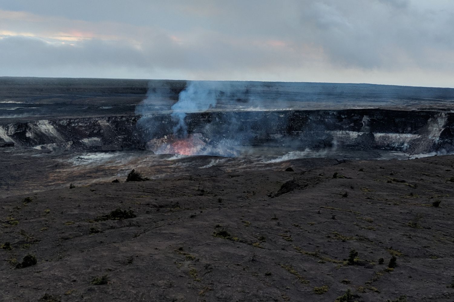 Dusk at Halemaʻumaʻu in January 2018: a red-orange glow from the lava lake deep in the crater wall, with blue volcanic fume rising over the dark crater floor.