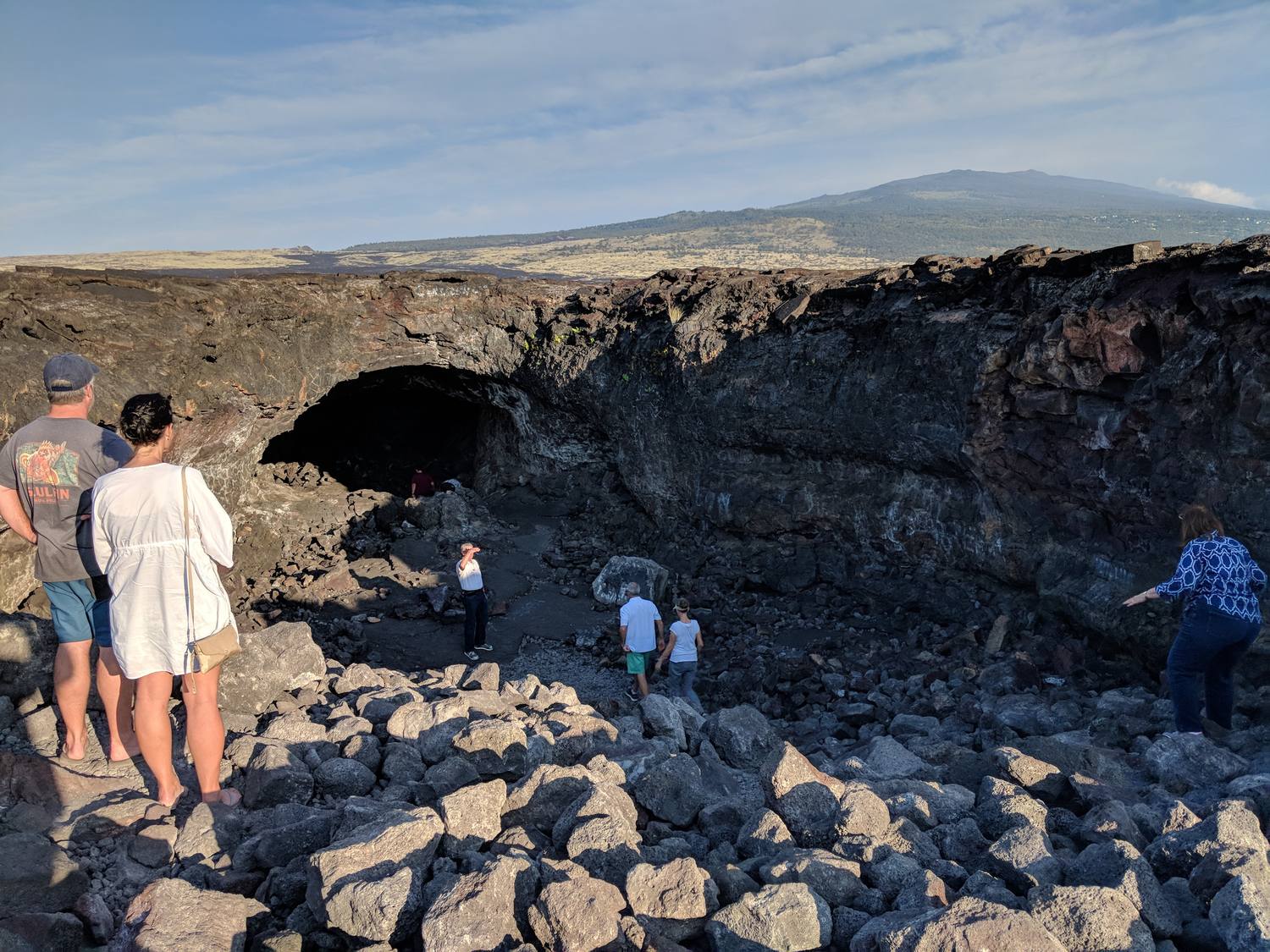 Family members climbing over jagged black lava boulders toward the dark mouth of a lava tube, with a broad green-and-brown volcano rising behind under a blue sky.