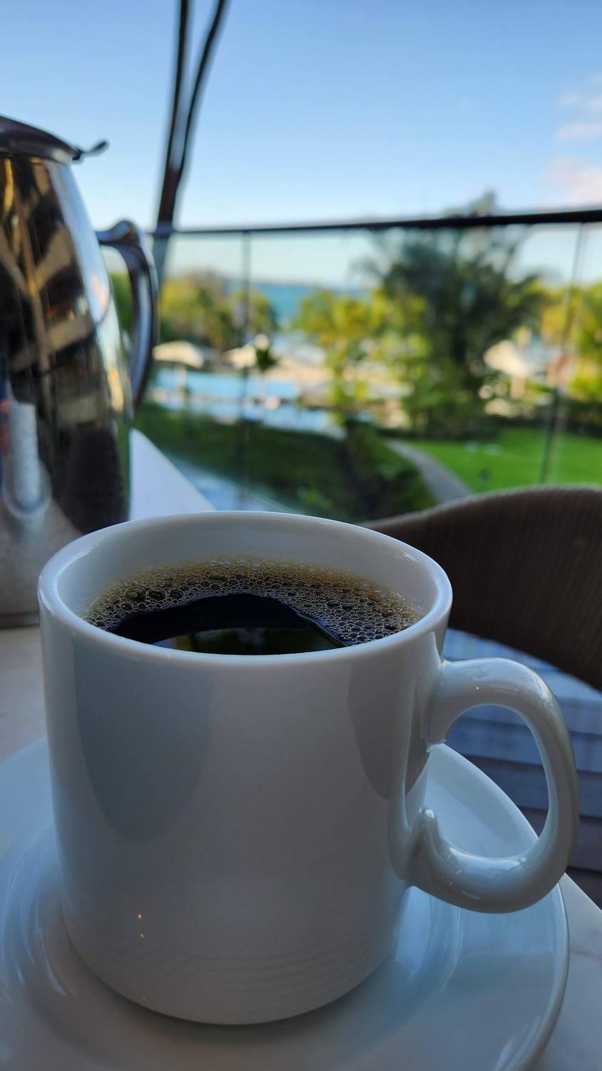 A white cup of black coffee on a saucer beside a silver coffee pot, on a terrace railing looking out over a pool, palms, and the ocean.