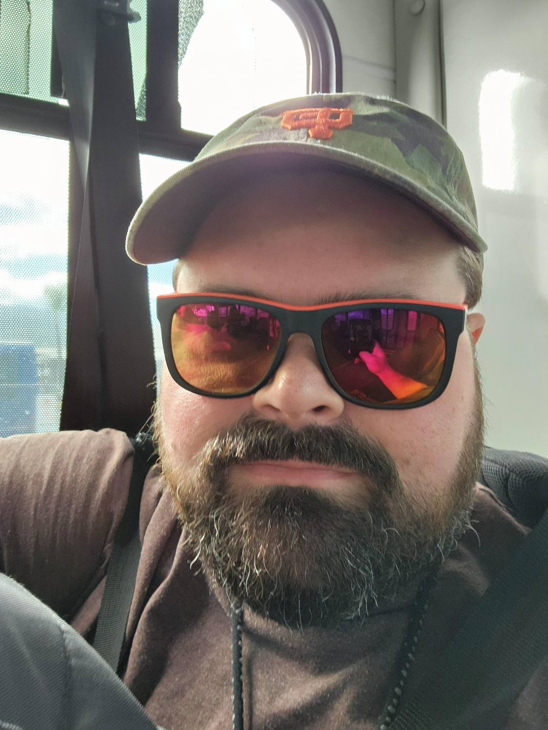 Joe Graham in a camouflage cap and red mirrored sunglasses, smiling, with a bus window behind him.