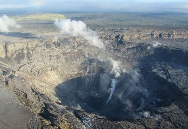 Aerial view of Halemaʻumaʻu after the 2018 collapse: a huge, deep, steaming pit with stepped cliff walls where the crater floor used to be.