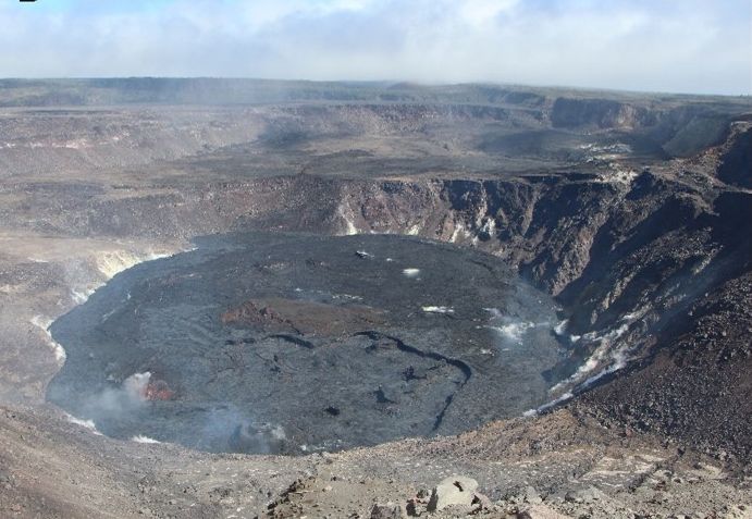 Halemaʻumaʻu after new eruptions: a wide, dark, solidified lava lake filling the bottom of the deep crater, with a small glowing vent at the edge.