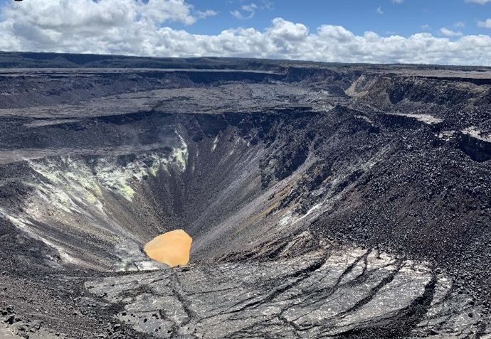 Halemaʻumaʻu in July 2019: a deep cone-shaped pit of gray rubble with a small orange-brown lake of water at the very bottom.
