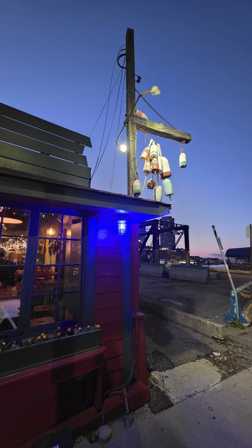 The red wooden Barking Crab restaurant at dusk, lit by a blue light, with a tall mast hung with buoys and a string of lights against a deep blue sky.
