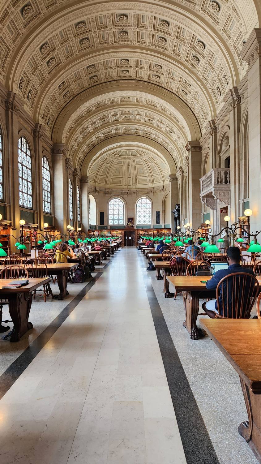 Bates Hall, a long reading room with a coffered barrel-vaulted ceiling, arched windows, and rows of wooden tables with green reading lamps, with people reading.