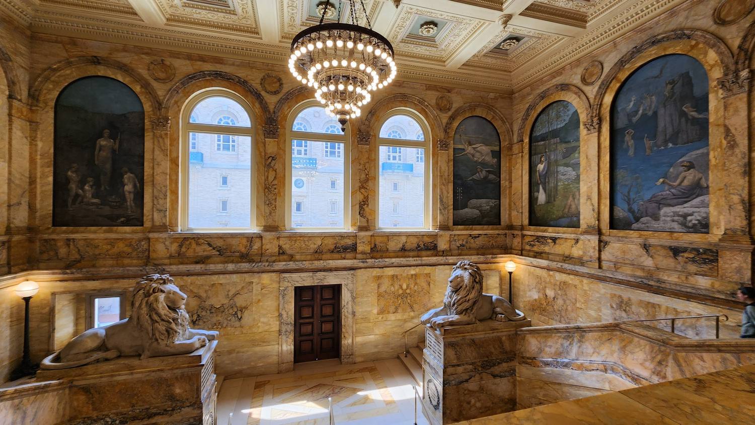The library's grand staircase of golden marble, with two marble lions on pedestals, a crystal chandelier, arched windows, and large pale blue murals on the walls.