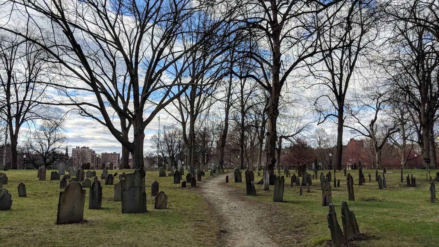 Rows of old slate headstones in green grass under tall bare trees, with a dirt path running through the middle and brick buildings beyond.