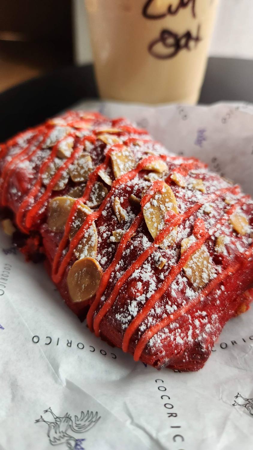 A bright red almond croissant striped with red icing and dusted with powdered sugar, on a bakery wrapper printed Cocorico, with a coffee cup behind it.