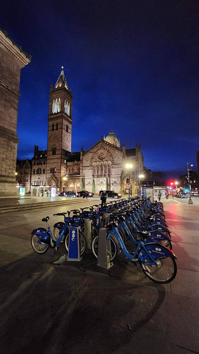 Copley Square at night: a row of blue rental bikes in a docking station in the foreground, and the tall stone bell tower of Old South Church lit against a deep blue sky.