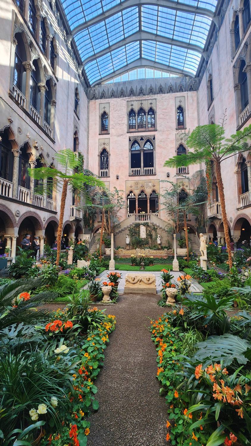 The Gardner Museum's glass-roofed courtyard, built like a Venetian palace, with pink walls, arched windows and balconies, tree ferns, statues, and beds of orange and purple flowers. Long orange nasturtium vines hang from balconies on both sides.