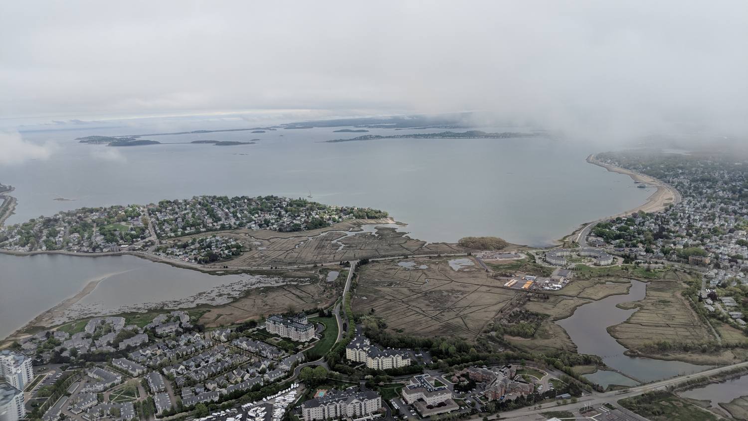 Looking down from a plane window at a gray bay, a peninsula of houses, salt marsh and a winding river in the foreground, a long curving beach on the right, and low islands on the hazy horizon.