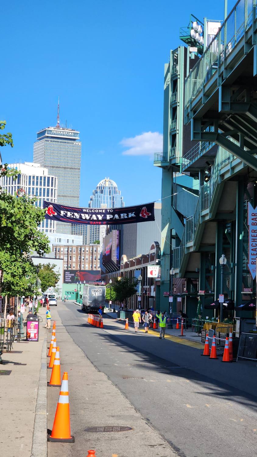 Jersey Street beside Fenway Park's green steel facade, with a Welcome to Fenway Park banner over the street, orange traffic cones, and the Prudential Tower rising in the distance under a blue sky.