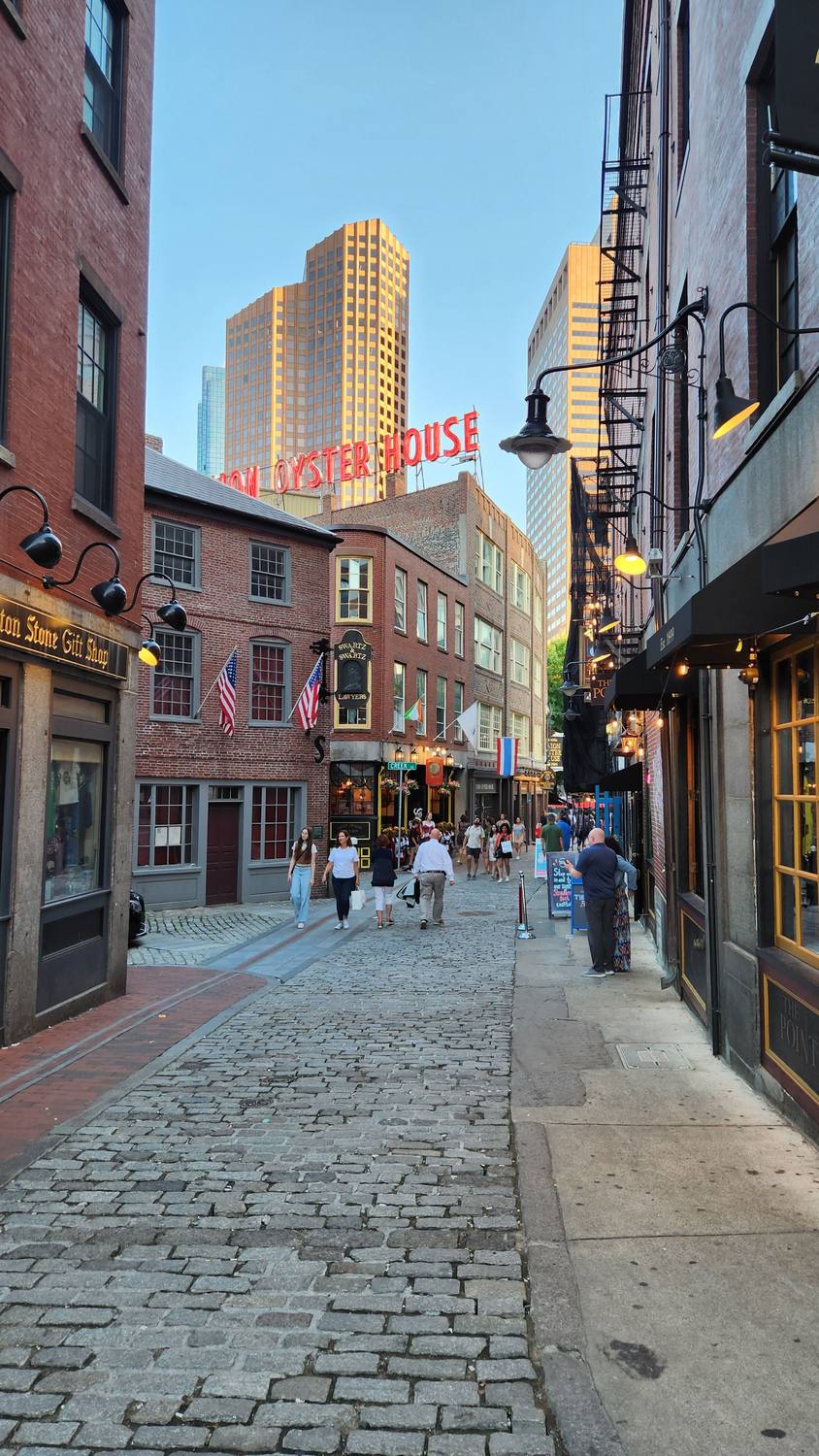 A narrow cobblestone lane between old brick buildings with gas-style lamps and American flags, leading to a red Union Oyster House sign, with modern towers rising behind it in evening light.