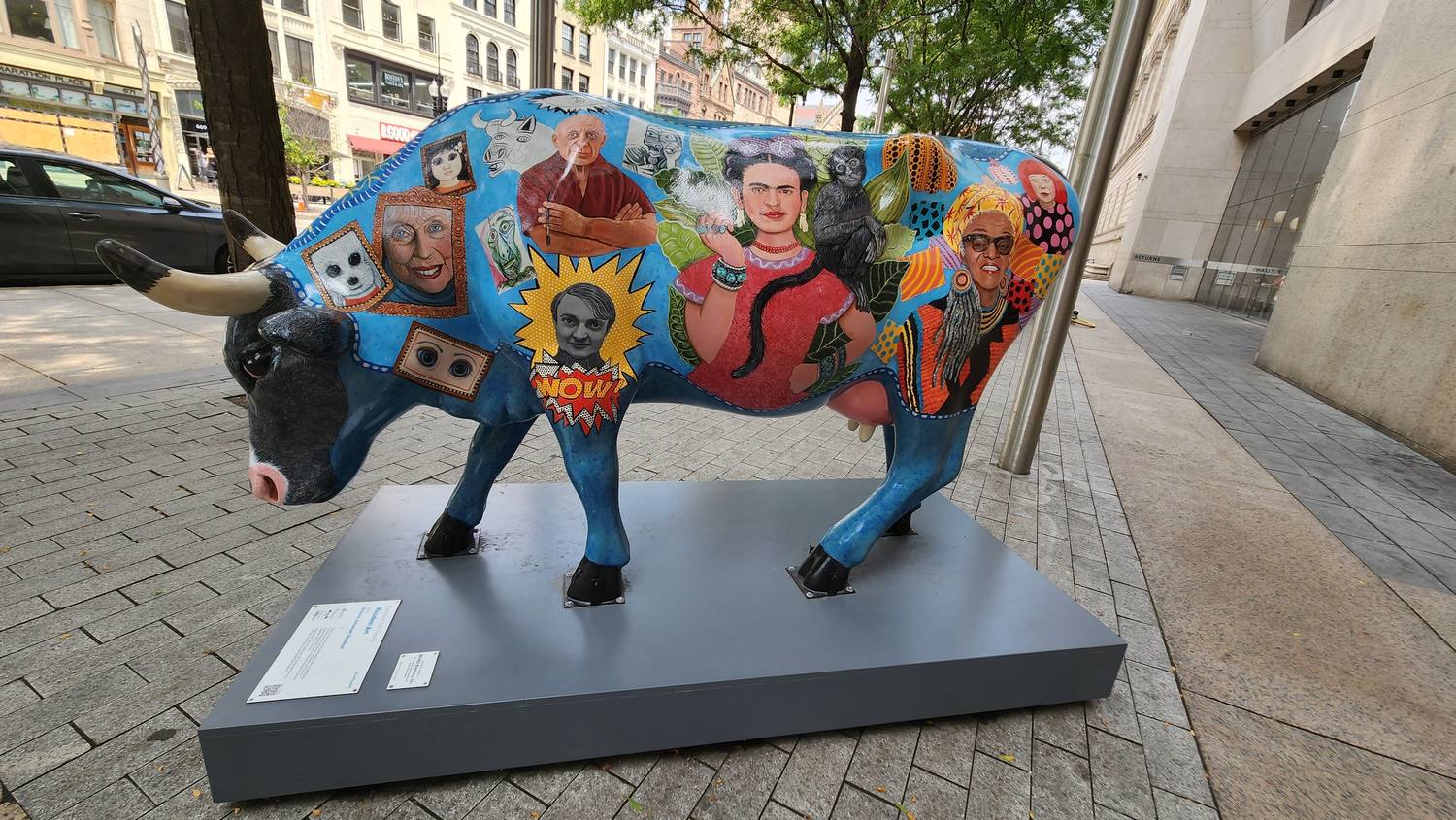 A life-size fiberglass cow painted blue and covered in portraits of artists, including Pablo Picasso, Frida Kahlo with a monkey, and Yayoi Kusama in polka dots, standing on a gray base on a brick sidewalk.