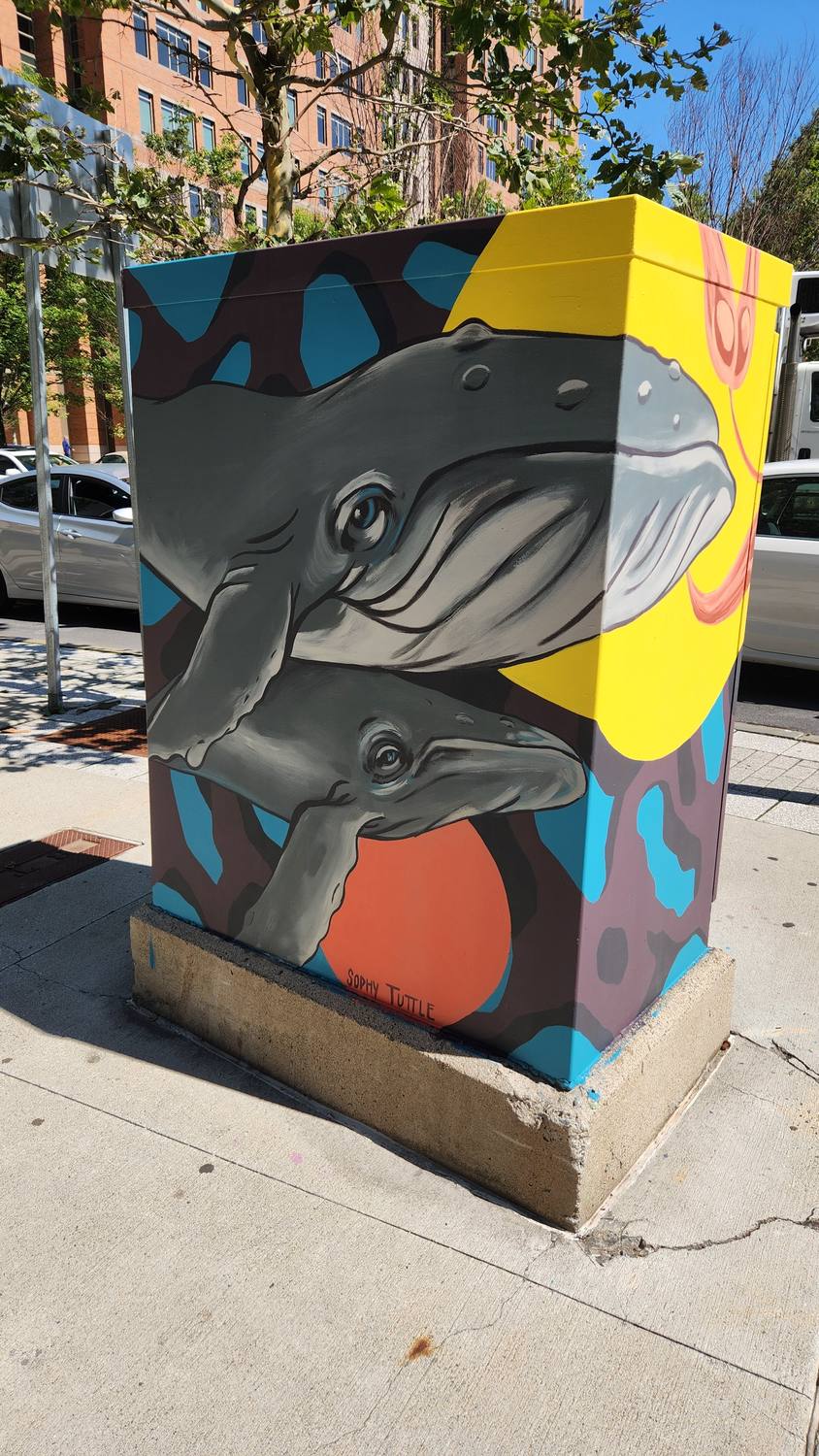 A sidewalk utility box painted with a gray humpback whale and her calf swimming across a yellow sun, over a pattern of blue, orange, and dark gray shapes.