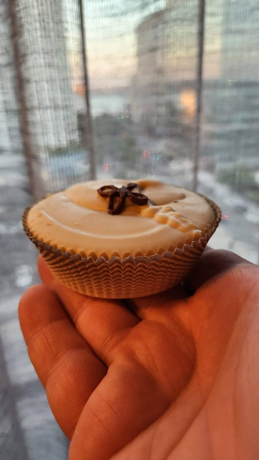 A hand holding a white chocolate candy cup topped with a small chocolate bow, in front of a blurry window view of city buildings.