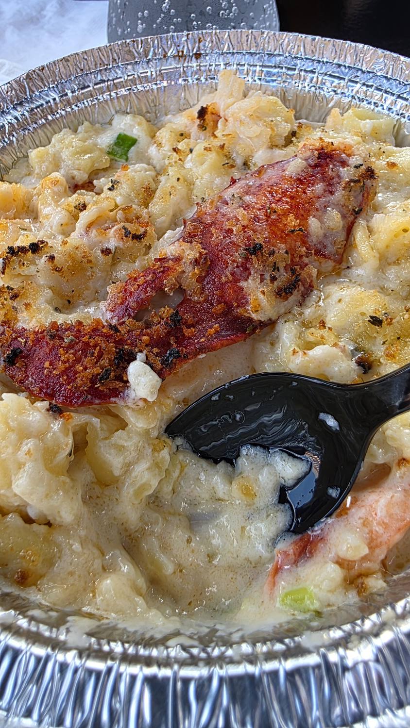 Lobster mac and cheese in a foil tin with a browned, crumb-topped crust, big pieces of lobster, and a black plastic spoon digging in.
