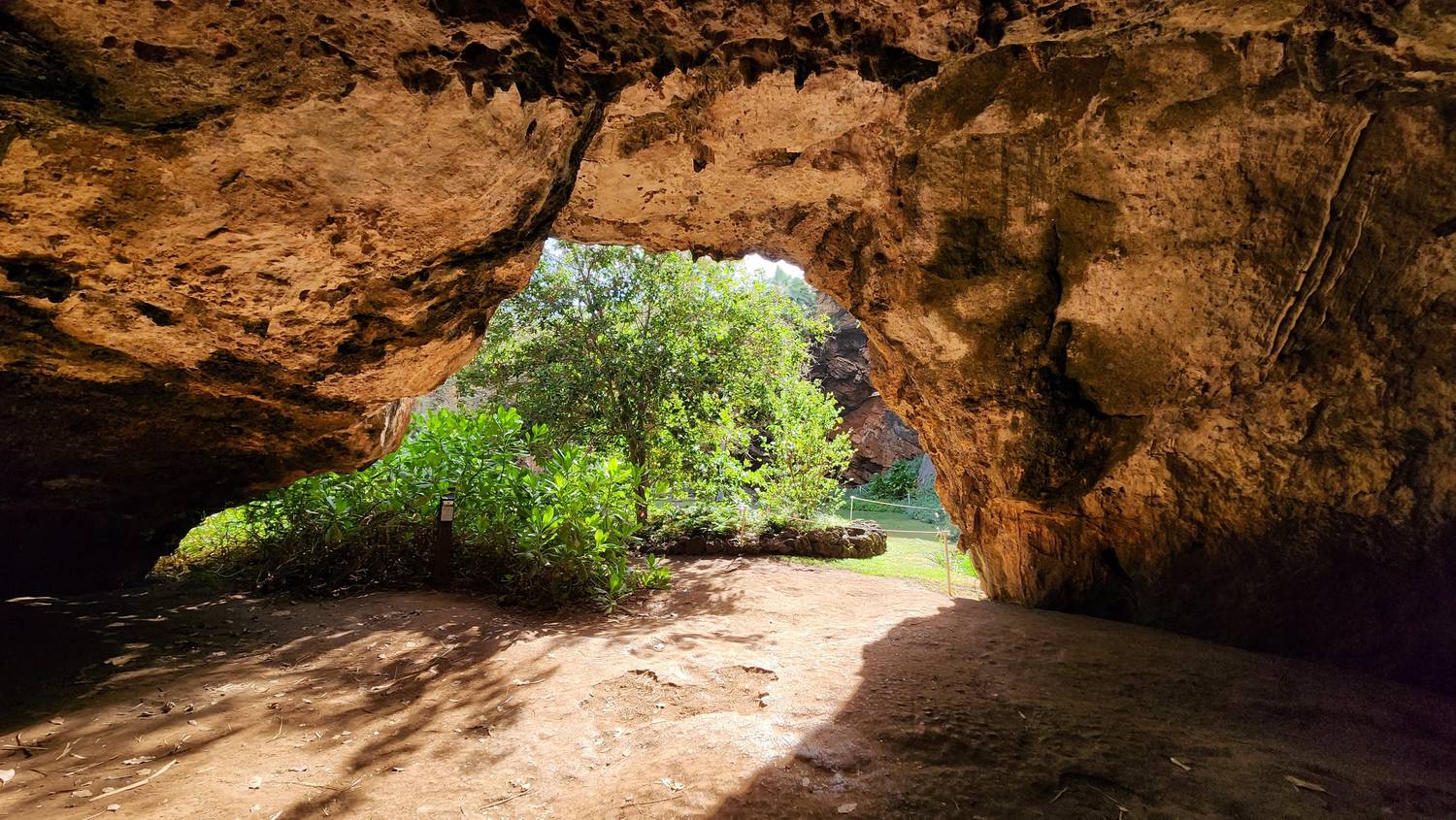 Inside the cave: a huge sculpted orange-brown limestone arch frames a bright opening full of green trees and shrubs, with a dirt floor in shadow.