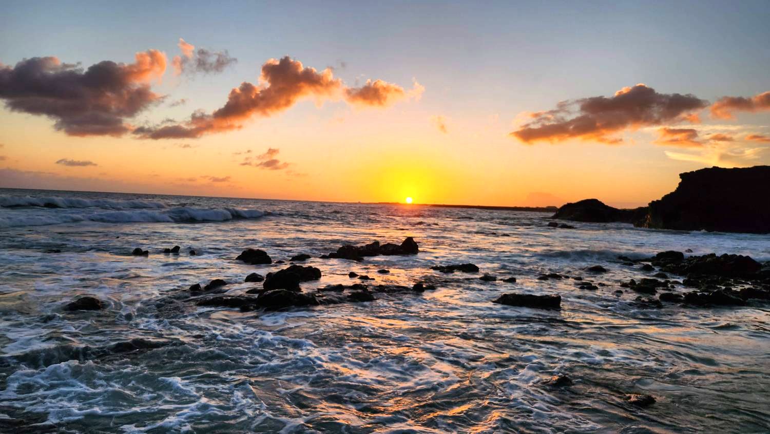 The sun setting on the horizon over the ocean, with pink and orange clouds, dark rocks in the foaming surf, and a dark headland on the right.