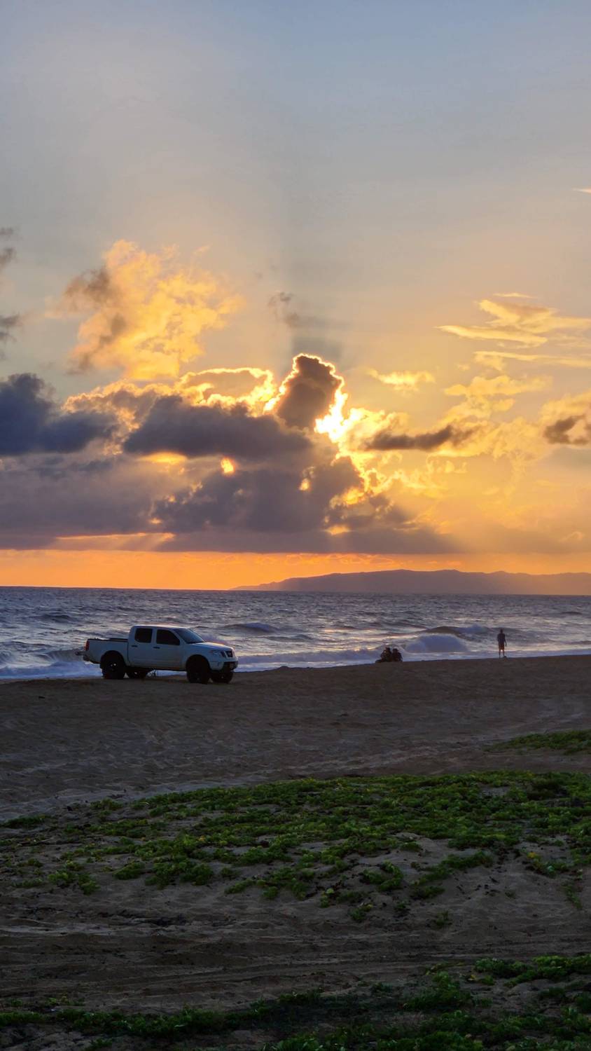 Sunset on the west side: sunbeams breaking through dark clouds over the ocean, a white pickup truck parked on the sand, and a long low island on the horizon.