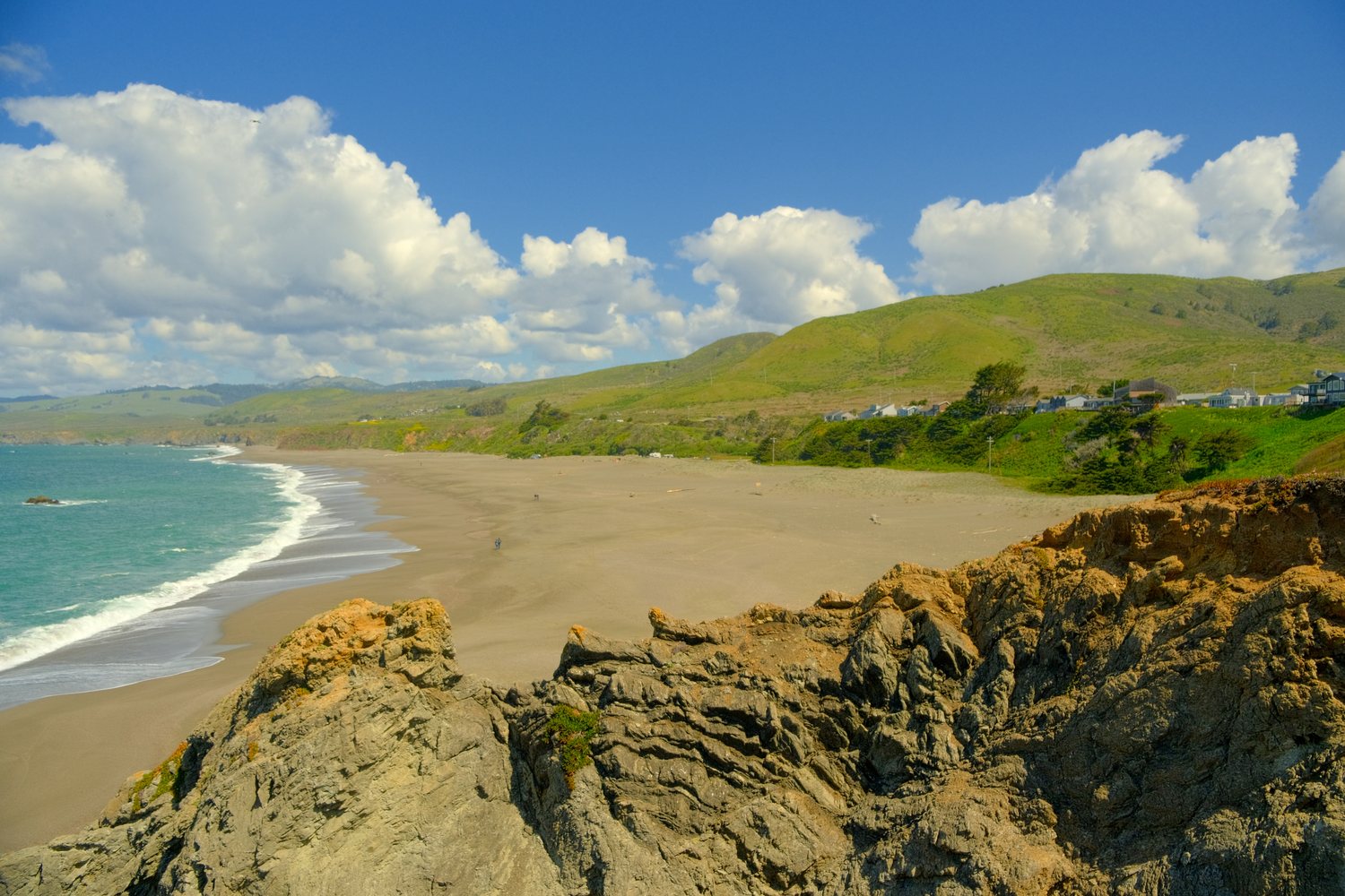 A long, wide sand beach curving toward green hills and a few houses, with surf on the left and rough rock in the foreground under big white clouds.