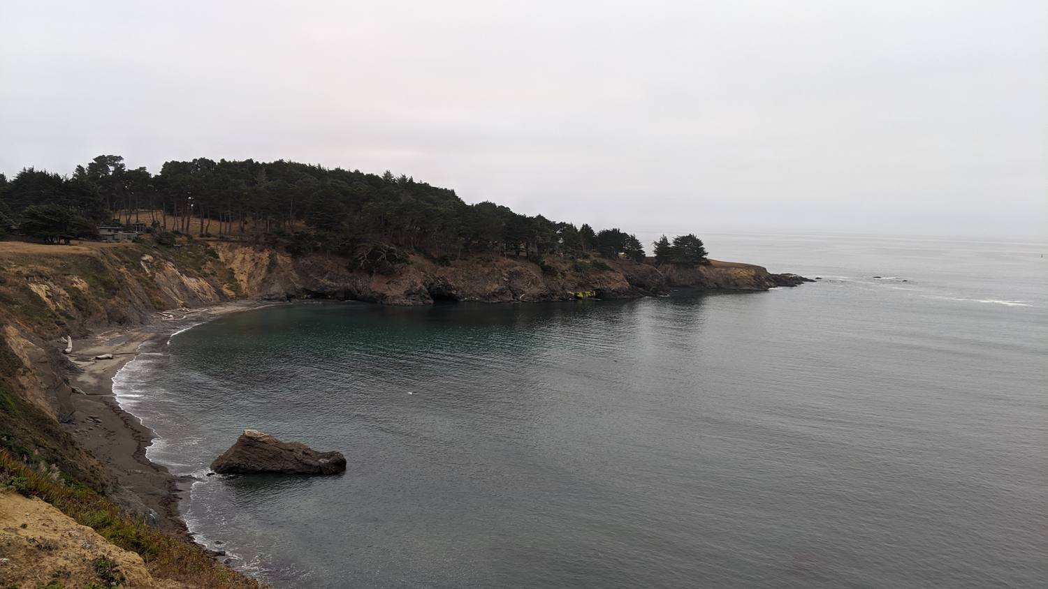 A calm gray cove under an overcast sky, ringed by eroded bluffs and a dark line of trees, with one large rock in the water near the beach.