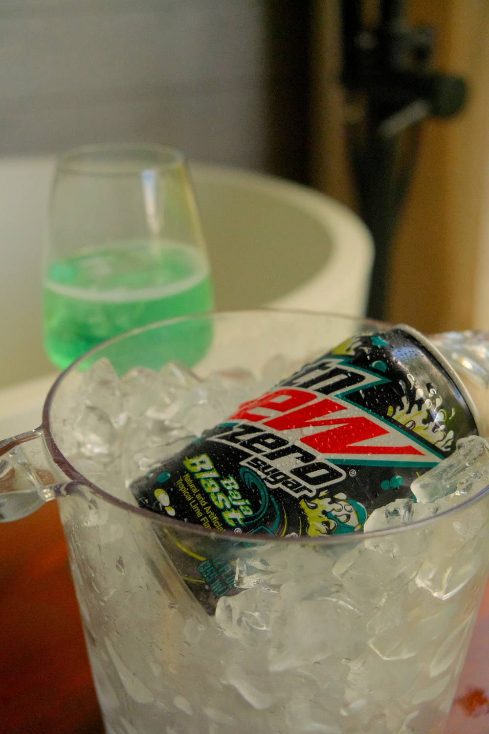 A can of Mountain Dew Zero Sugar chilling in a clear ice bucket, with a glass of bright green soda on the table behind it.