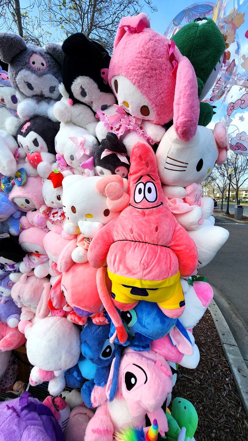 A tall wall of stuffed toys, including Hello Kitty, Kuromi, My Melody, and a pink Patrick Star, piled against a blue sky.